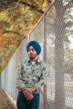 男子身着图案衬衫，头戴头巾，倚靠在旁边的金属围栏上，被捕捉于户外场景中