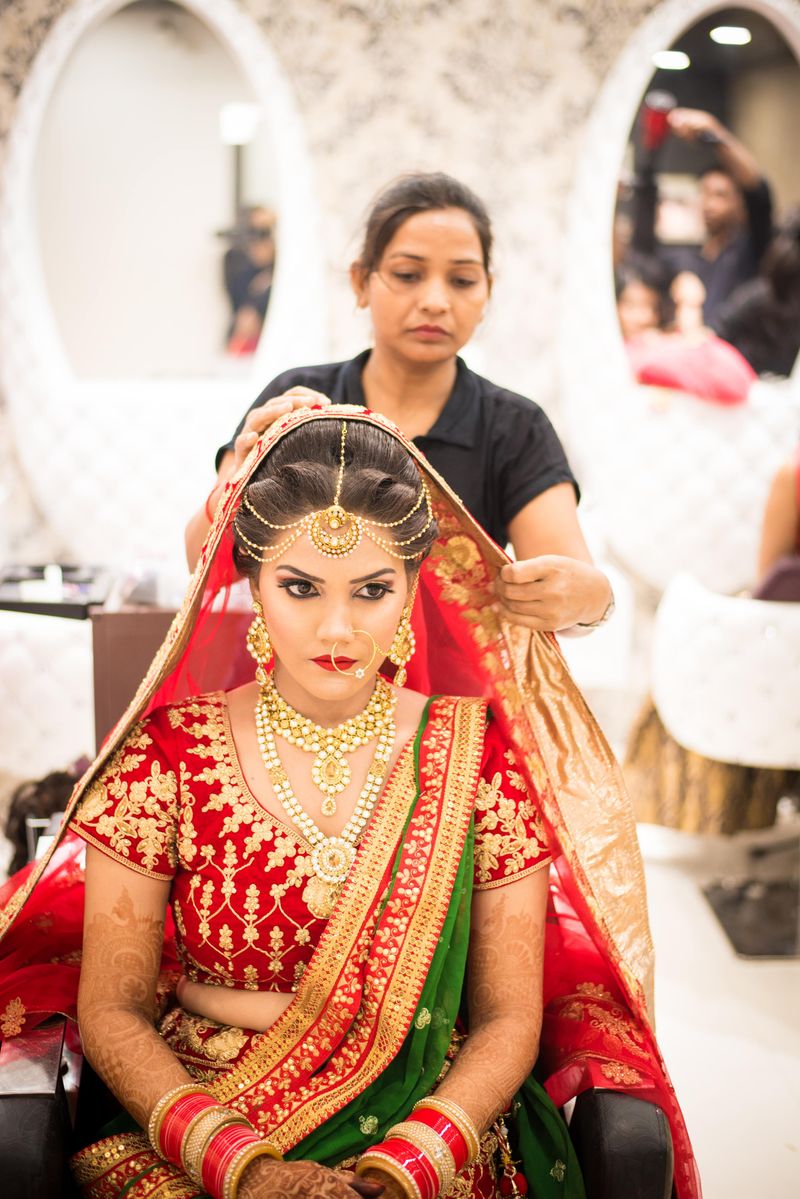 A bride in red and gold having her dupatta set by a stylist before the ceremony.