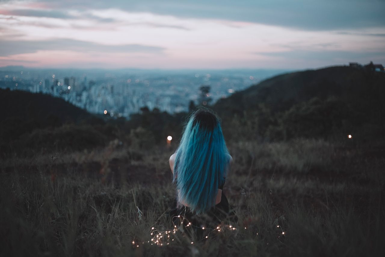 a woman with blue hair sits on a hillside gazing at the city s twiligh