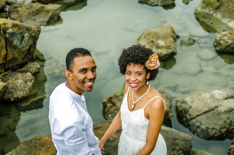 Photo Of Smiling Couple Dressed In White Holding Hands While Standing On Rocks By A Body Of Water