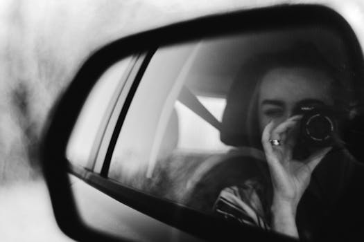 Black and white close-up of a woman taking a photo through a car's side mirror.