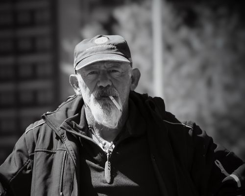 Free Grayscale portrait of an elderly man with a beard, wearing a cap and jacket, smoking outdoors. Stock Photo