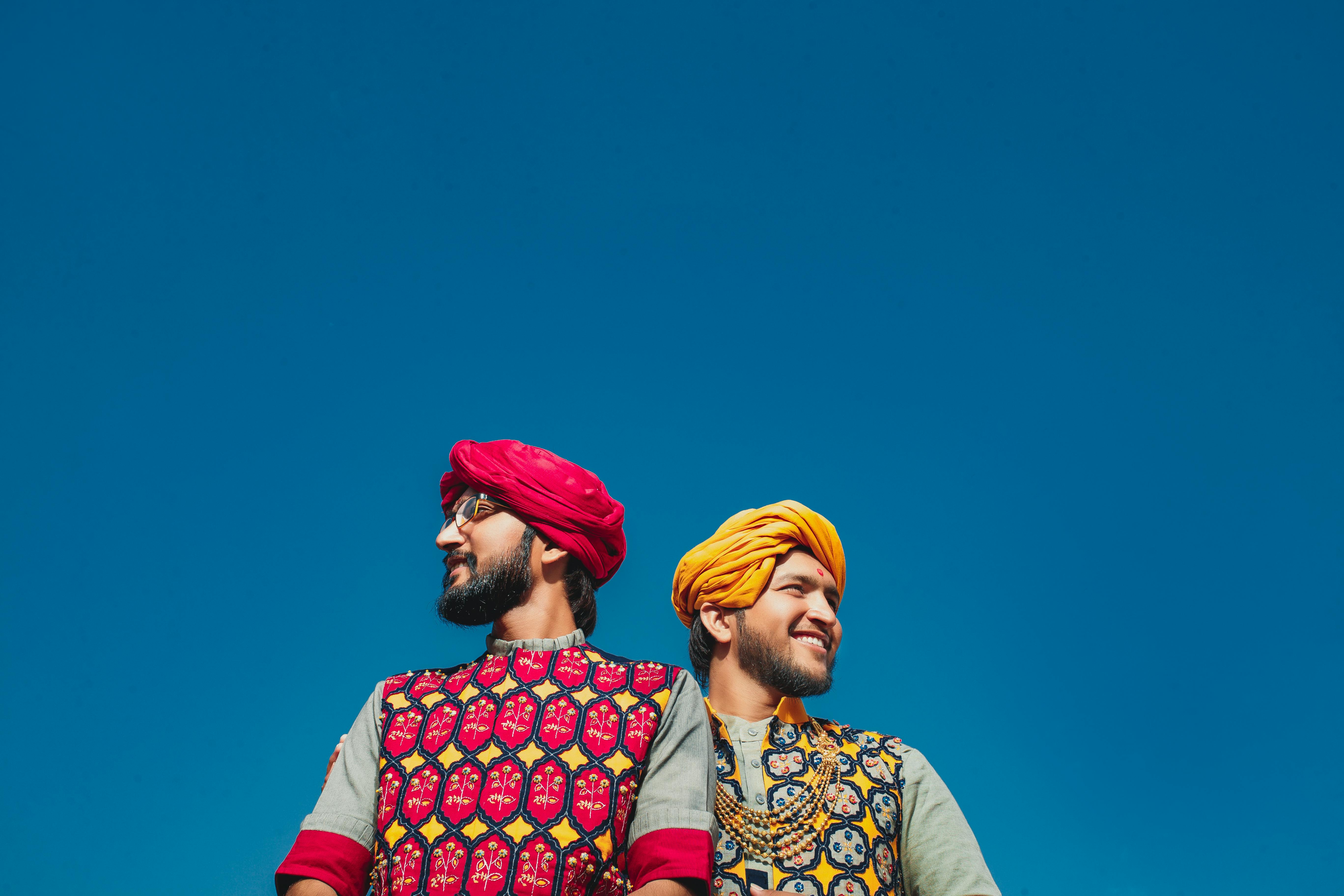 Two men in vibrant traditional attire looking in opposite directions under a clear blue sky.