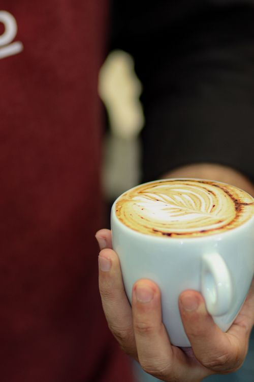 Free Hand holding a cappuccino with intricate latte art in a cozy cafe setting. Stock Photo