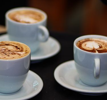 Three cappuccinos with intricate latte art in white cups on saucers, showcasing coffee artistry.