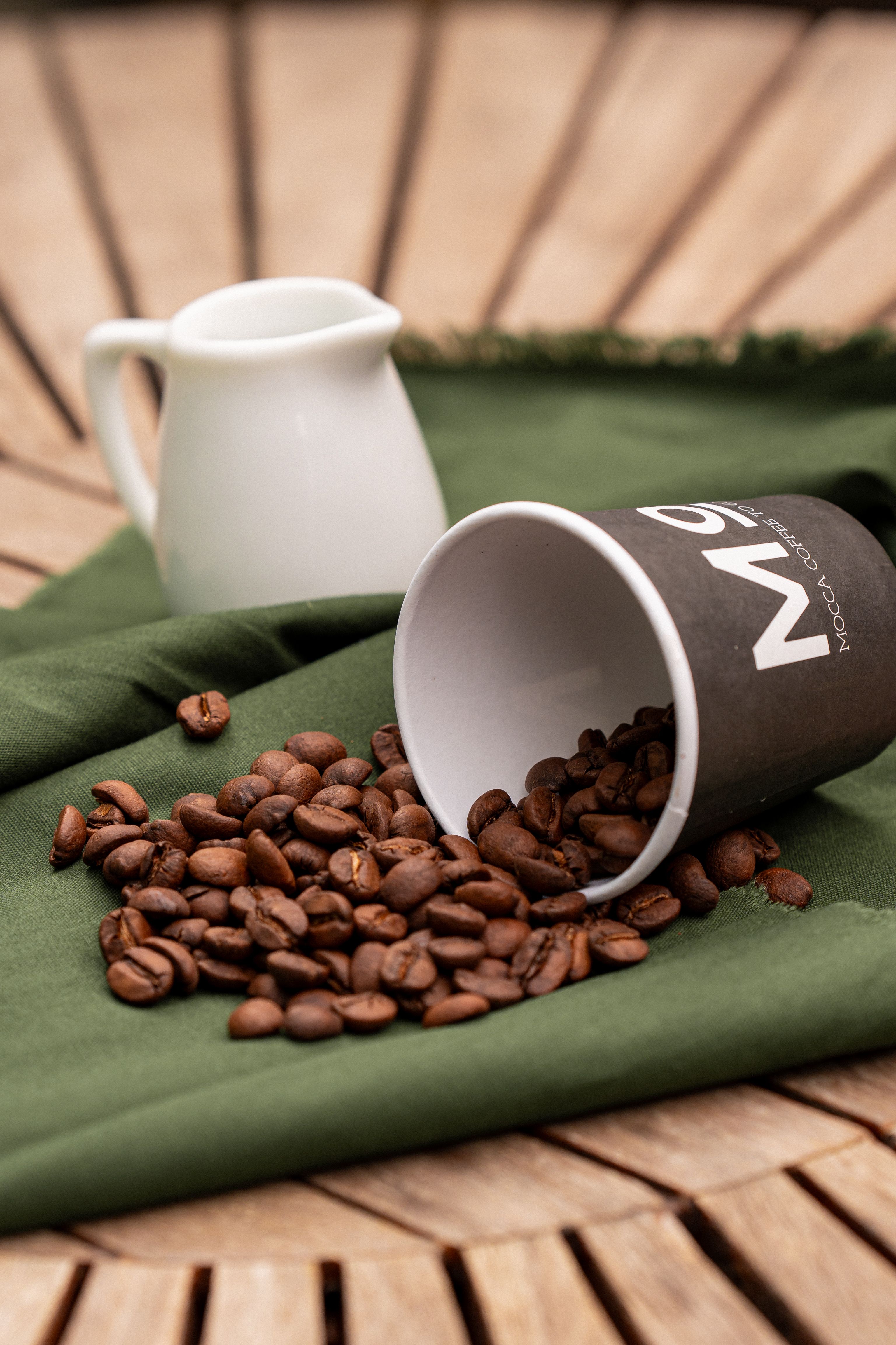 Close-up of coffee beans spilling from a cup onto a green cloth with a milk jug in the background.