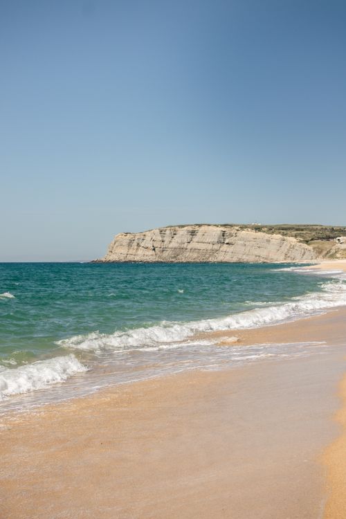 Free Peaceful beach with waves crashing onto sandy shore and cliffs under clear blue sky. Stock Photo