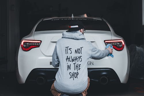 A man kneeling down in front of a car with a hoodie that says it's not always in the shop