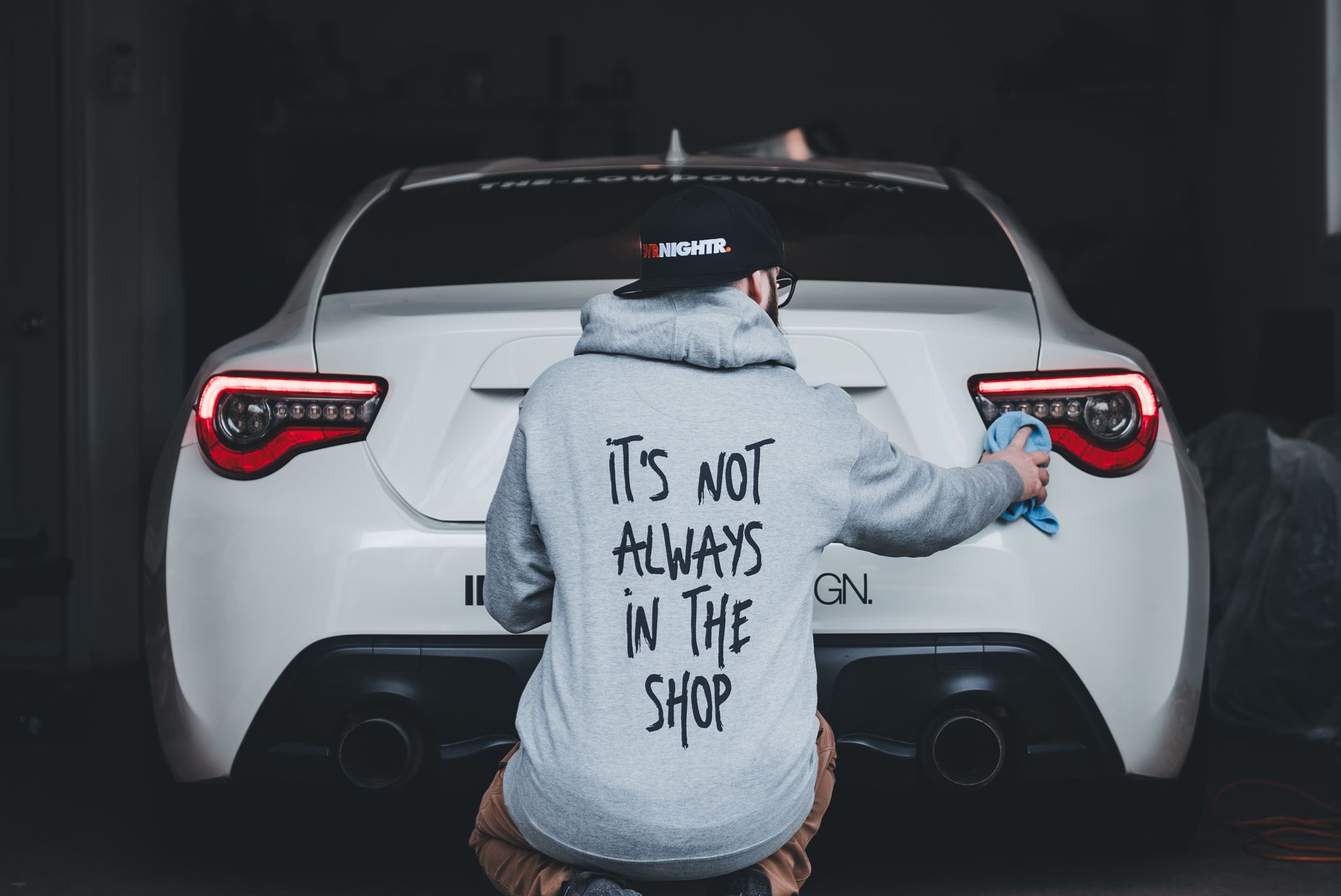 Man in hoodie cleaning a white sports car in a garage. Text on hoodie says 'It's Not Always in the Shop.'