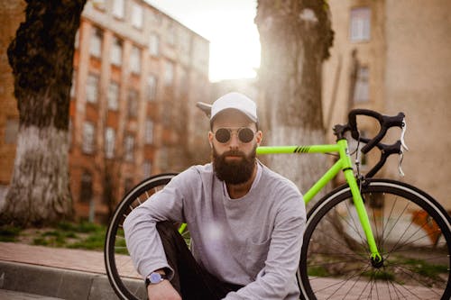 Bearded male in trendy outfit and sunglasses sitting near bike and looking at camera while resting on city street