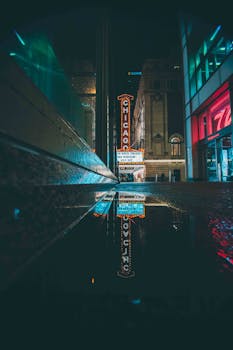 Chicago theater sign reflected in puddle amidst urban cityscape at night.