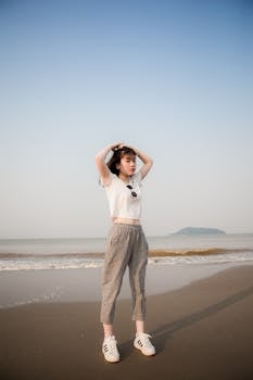 一位穿着露脐上衣和格子裤的年轻女子，在阳光明媚的海滩上对着安详的海浪摆姿势