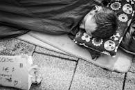 一张黑白照片里，一名无家可归者睡在城市人行道上，旁边有一个标志和一些硬币