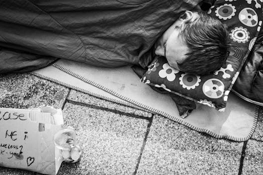 一张黑白照片里，一名无家可归者睡在城市人行道上，旁边有一个标志和一些硬币