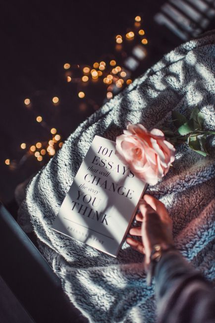 Hand reaching for a book and rose on a cozy bed, lit by warm fairy lights.