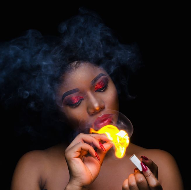 Portrait of a woman with striking makeup lighting a match, creating dramatic smoke and flames.