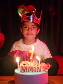 Young boy wearing crown seated behind birthday cake with candle. Joyful indoor party ambiance.