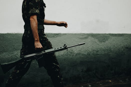 A soldier in camouflage walking with a rifle against a textured wall, day.