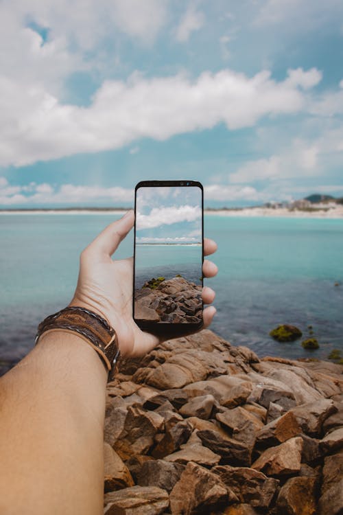 Crop anonymous traveler using mobile phone while taking shot of amazing scenery of rocky sea shore during summer holidays