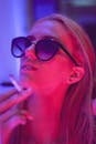 霓虹灯下，一个戴着墨镜的时尚女人在吸烟的特写肖像