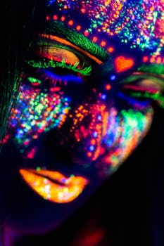 Close-up of a woman's face painted in neon colors under UV light, creating a dramatic visual effect.