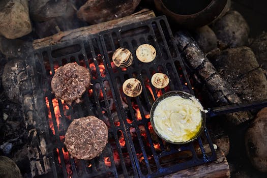 Juicy grilled meat patties with onions and sauce over a campfire in nature.