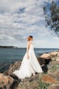 身着优雅白色礼服的新娘站在海边的岩石上，晴朗的天空下凝视地平线