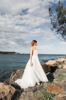 身着优雅白色礼服的新娘站在海边岩石上，在晴朗的天空下凝视着地平线