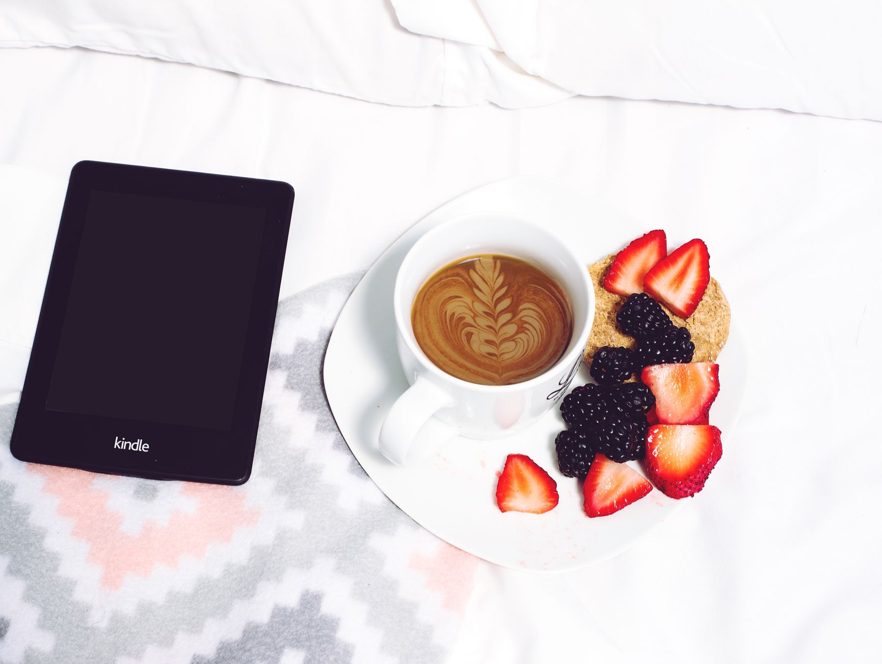 Relaxing morning setup with coffee, berries, and Kindle on a bed, offering a perfect cozy vibe.