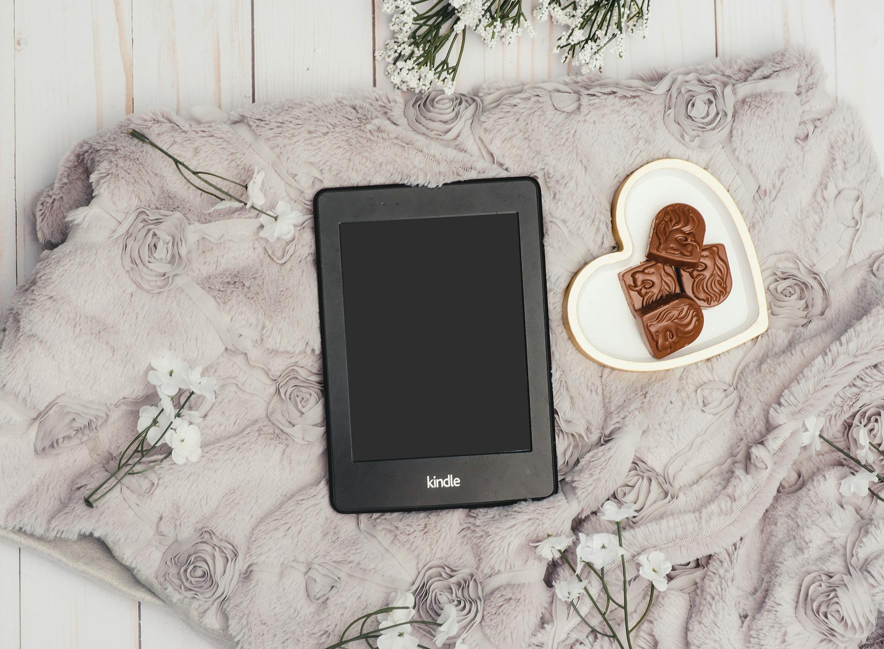 A Kindle on a blanket with heart-shaped chocolates and flowers for a cozy reading ambiance.