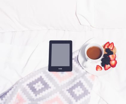 Flat lay of a Kindle, coffee, and berries on a cozy bed setting, perfect for relaxation.