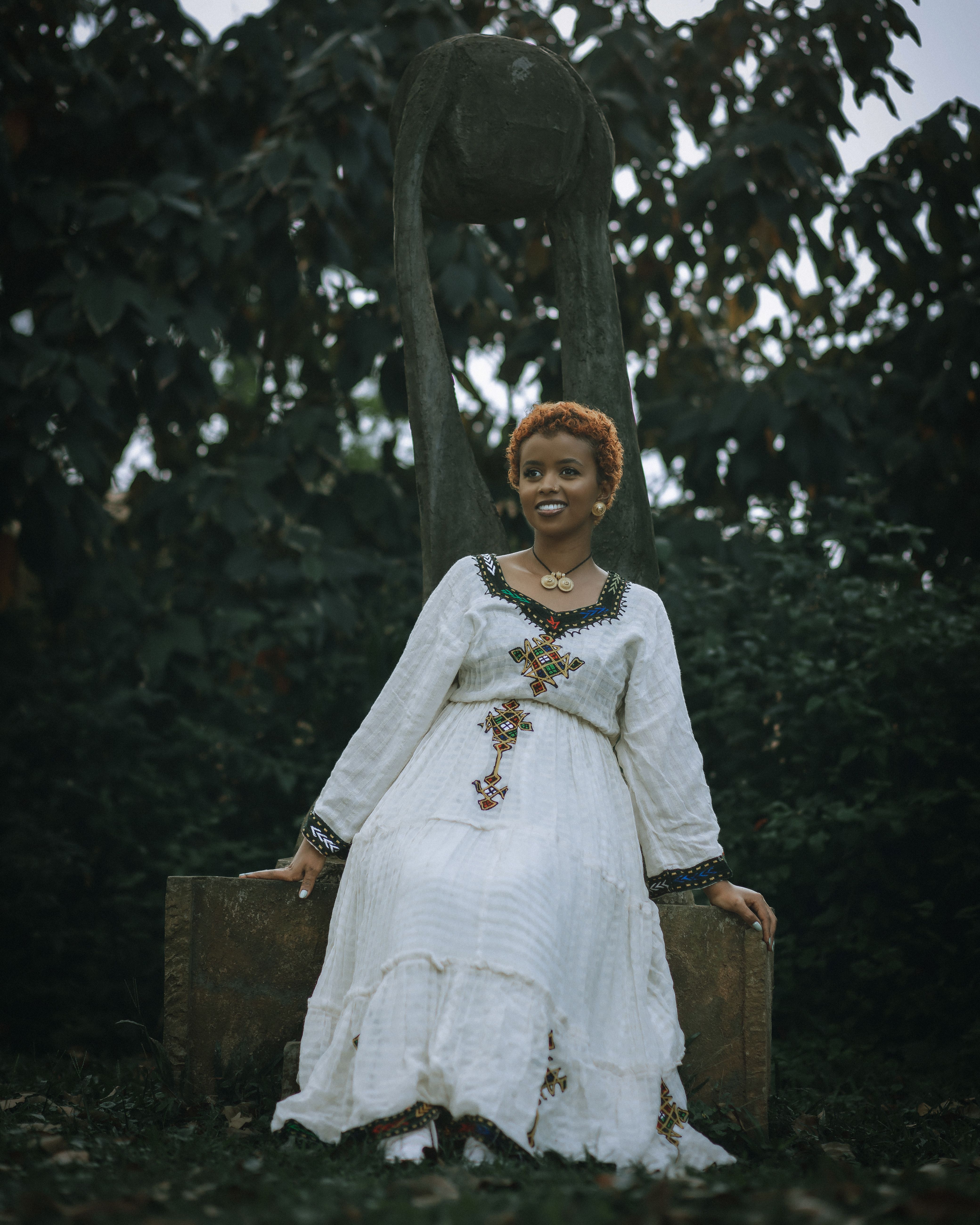 Woman sitting in elegant white dress with embroidery, posing outdoors against a lush park backdrop.