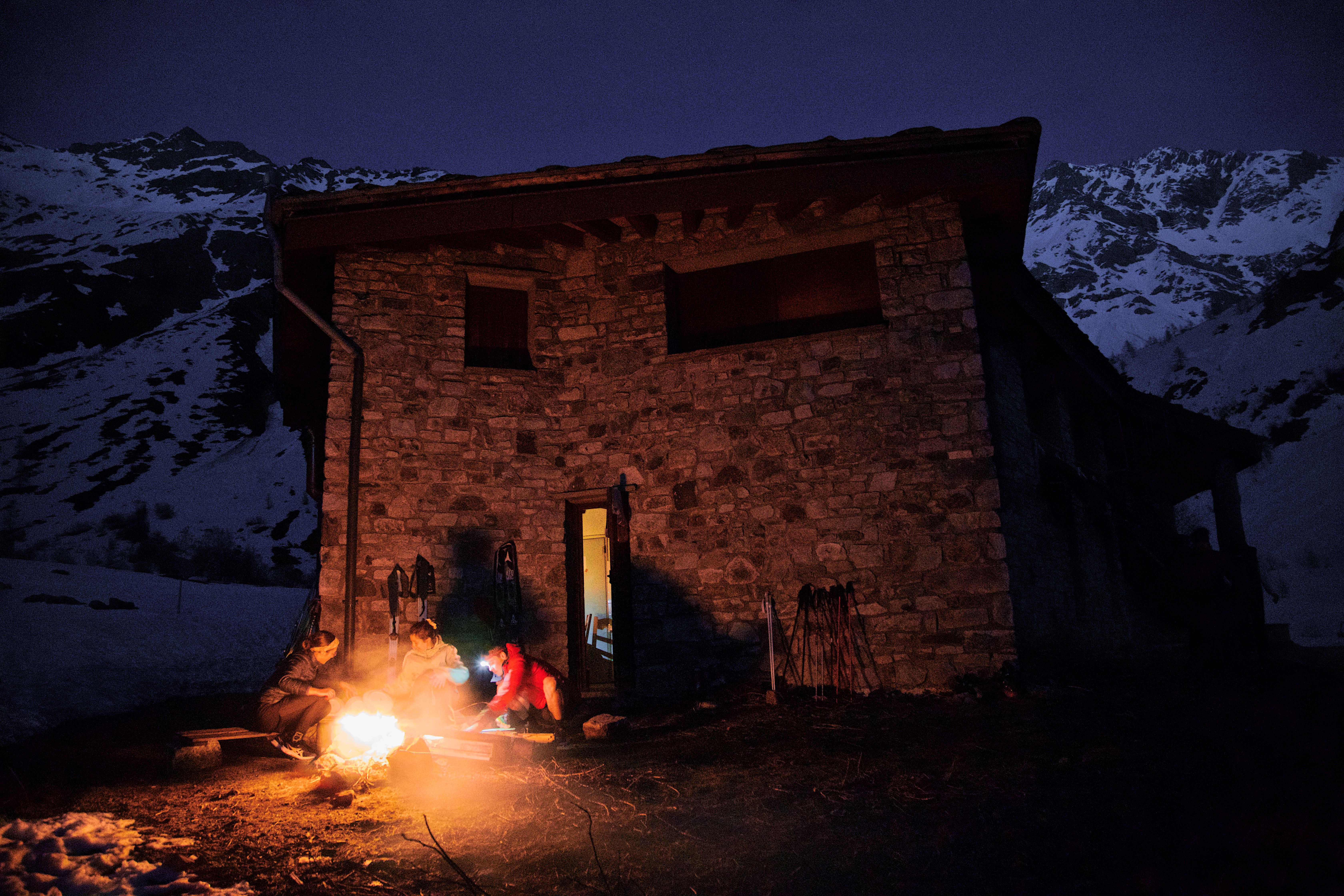 Rustic mountain cabin with a campfire at dusk, surrounded by snow-capped peaks.