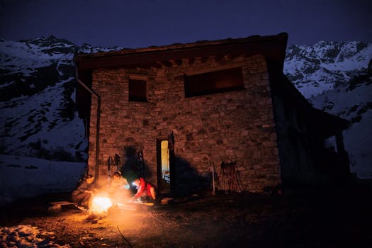 Rustic mountain cabin with a campfire at dusk, surrounded by snow-capped peaks.