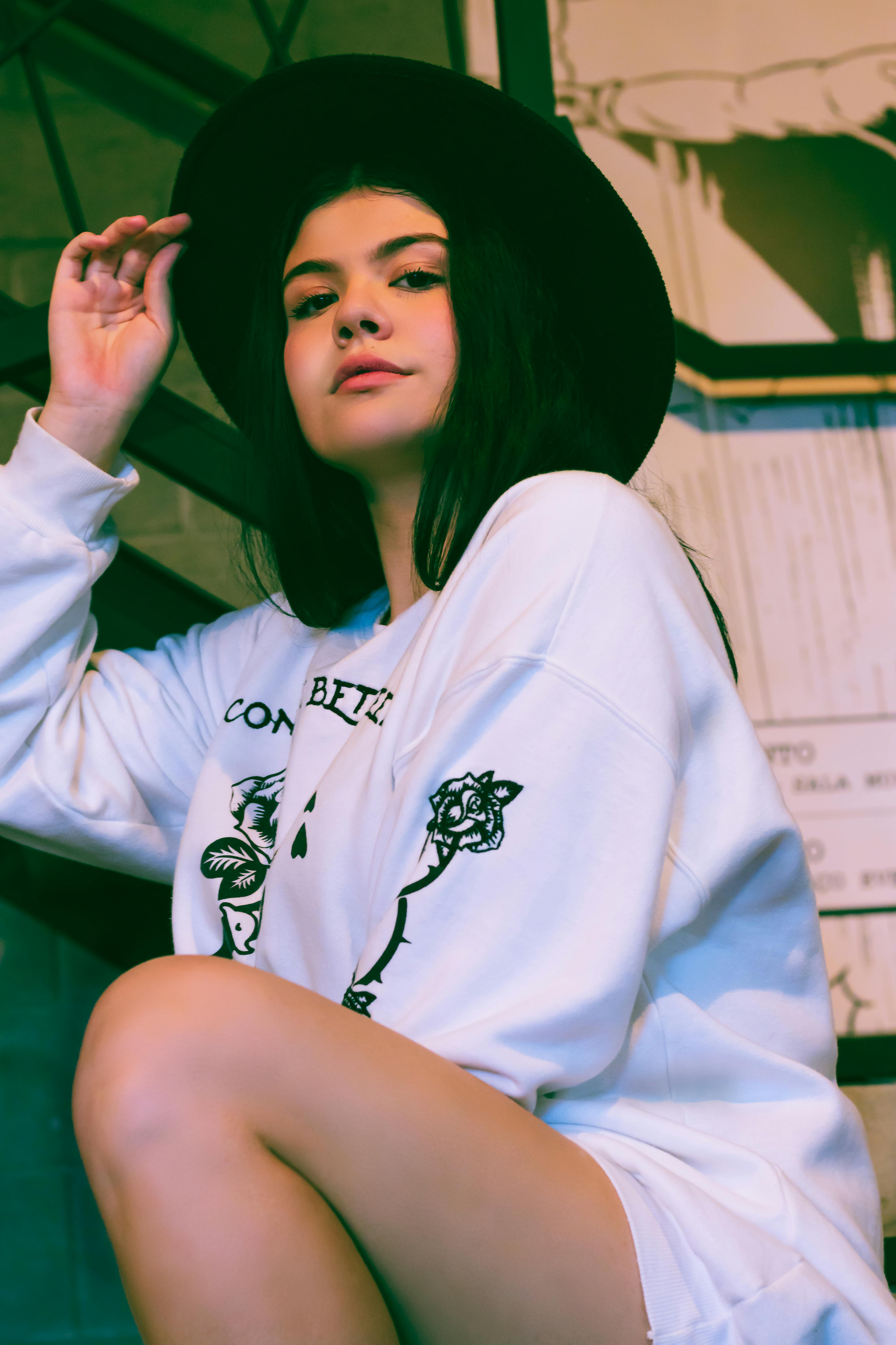 A fashionable woman in casual wear and a hat posing confidently indoors.