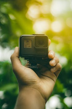 Close-up of a hand holding an action camera against a green leafy background.