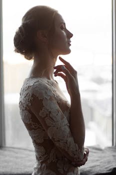 一位穿着蕾丝礼服的新娘优雅地靠在背光的窗户上摆出姿势