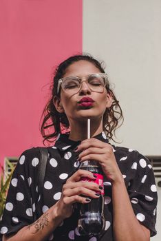 一位戴着眼镜、穿着圆点连衣裙的时尚女性，正靠着户外的彩色墙壁喝饮料