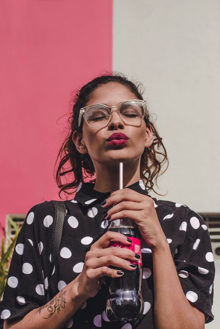 Trendy woman in polka dot dress wearing glasses, sipping a beverage outdoors against colorful wall.