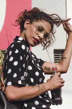 Trendy woman in polka dot shirt adjusting hair while holding soda bottle, outdoors.