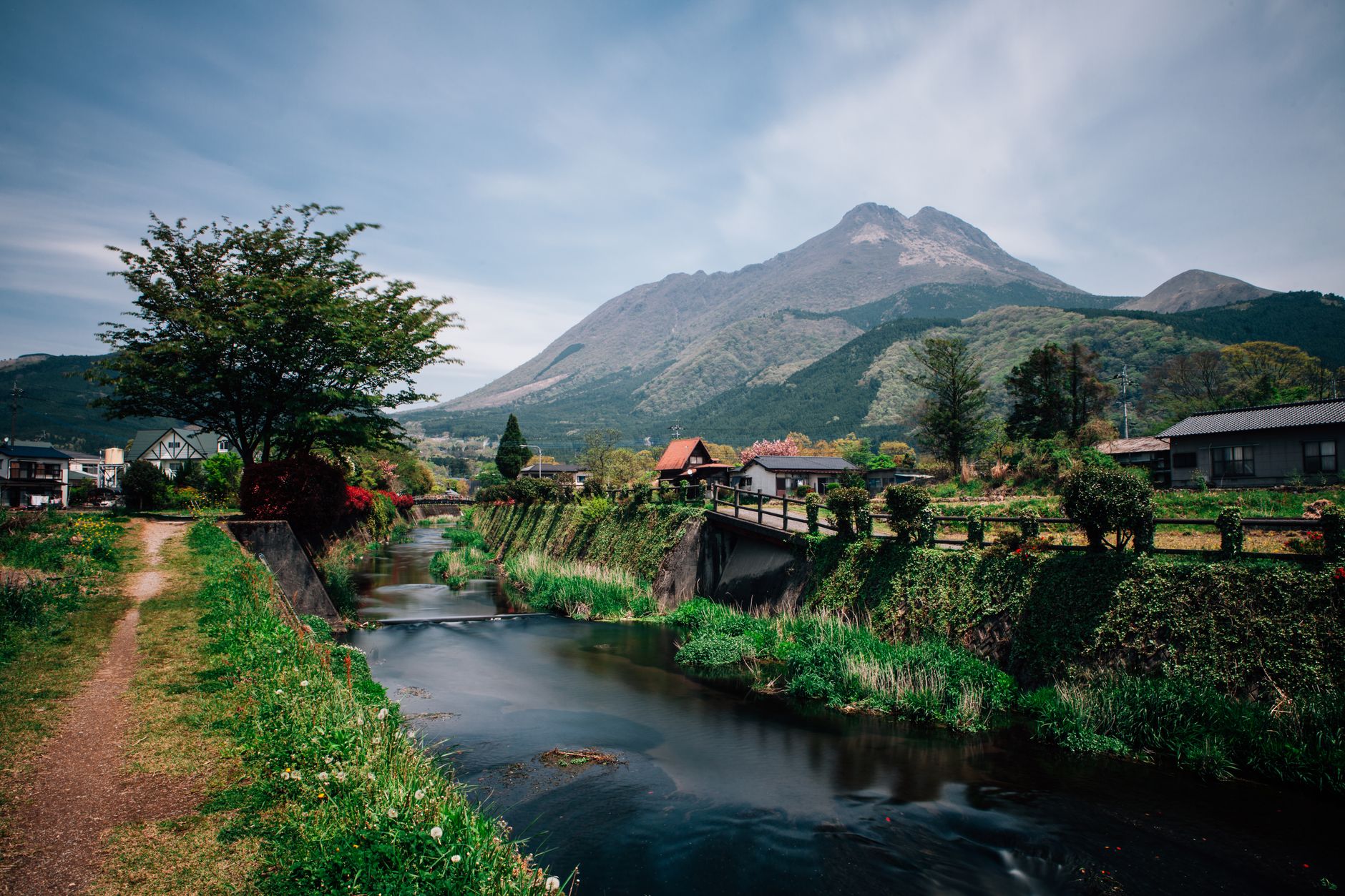 Charming mountain village landscape featuring a serene river, lush greenery, and a majestic mountain backdrop.