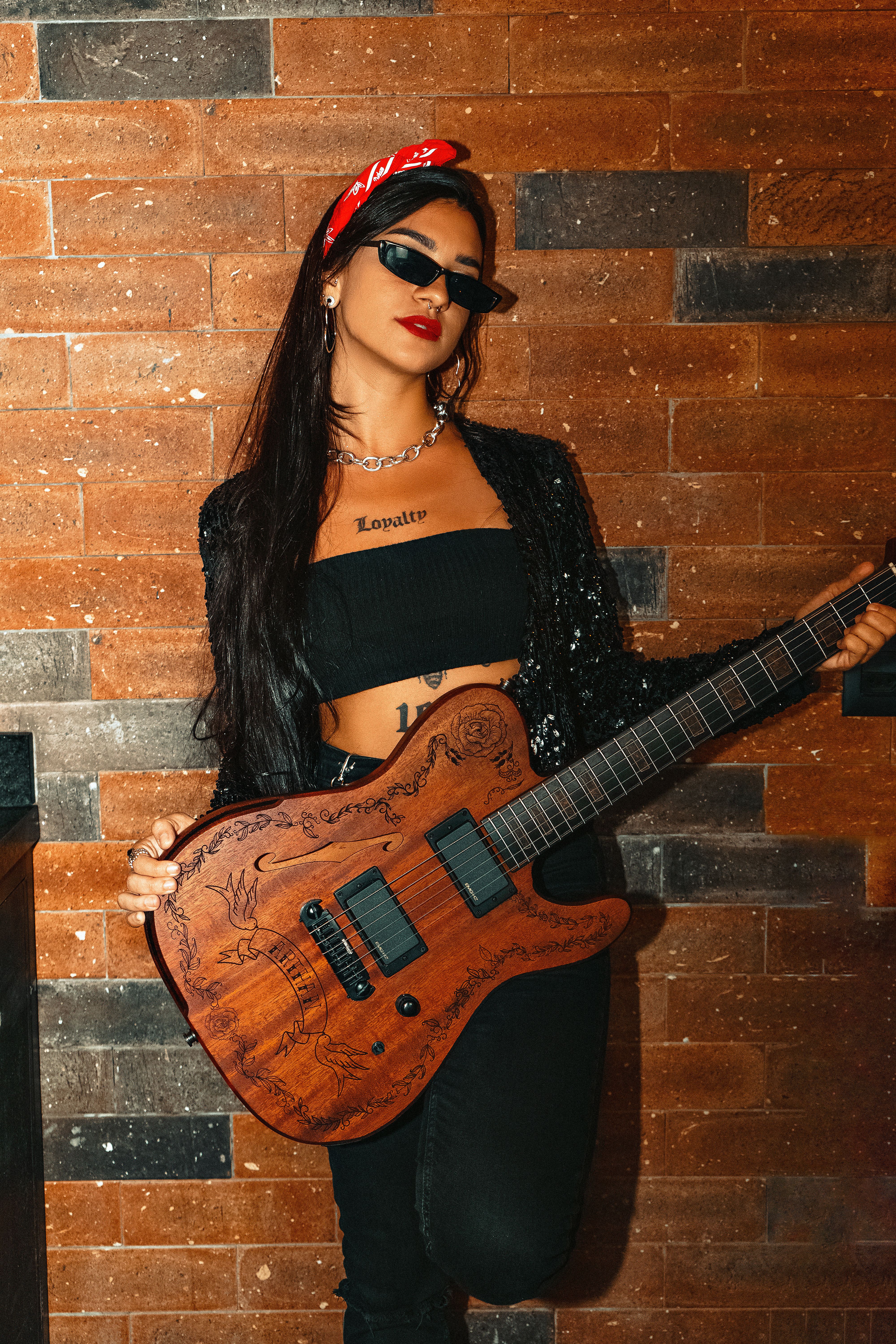 Trendy woman with sunglasses and guitar against rustic wall, exuding cool confidence.