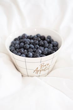 A bowl of fresh blueberries in a stylish "Bon Appétit" bowl, placed on a soft white surface.