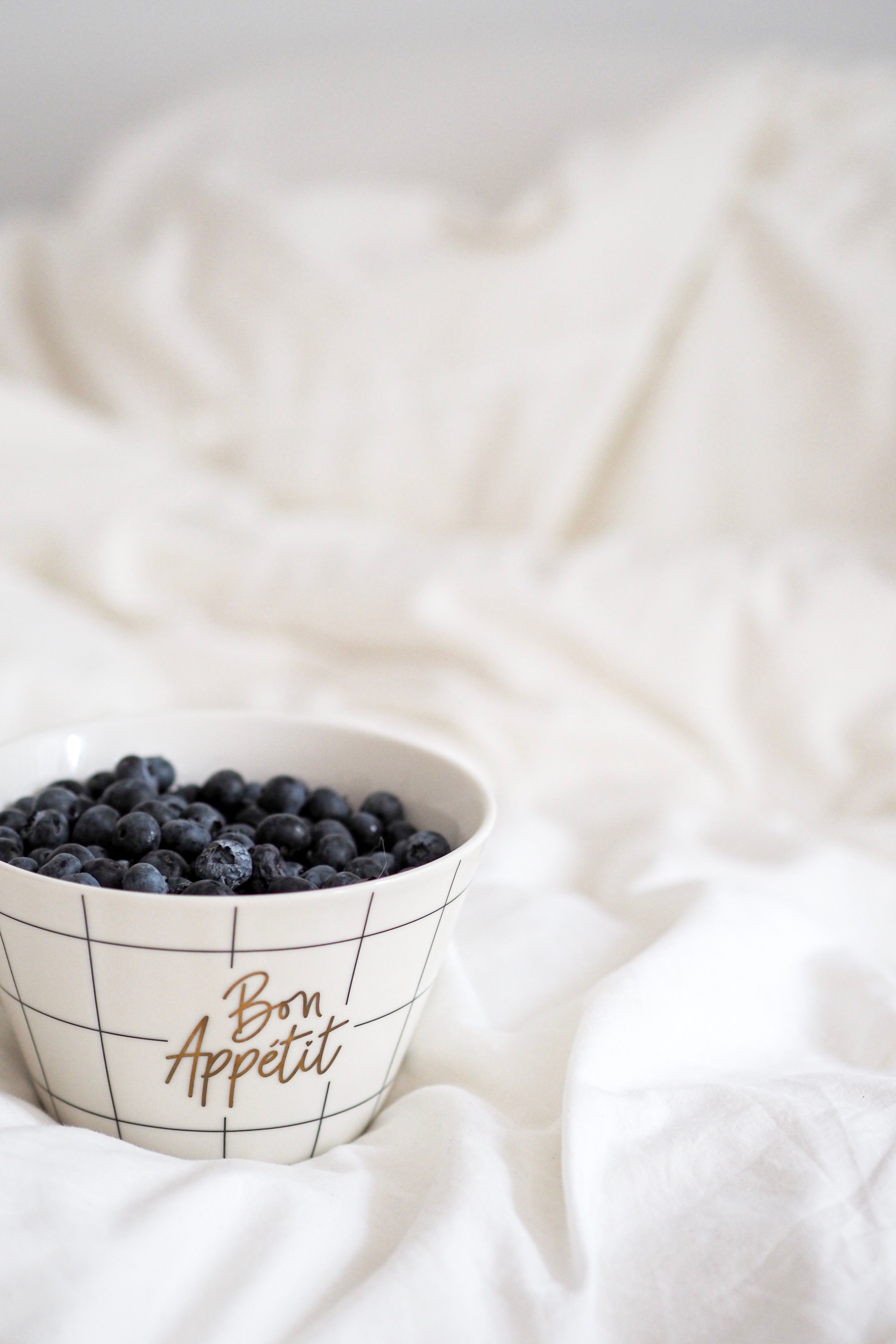 High angle shot of blueberries in a ceramic bowl with 'Bon Appetit' text, placed on a soft white cloth.