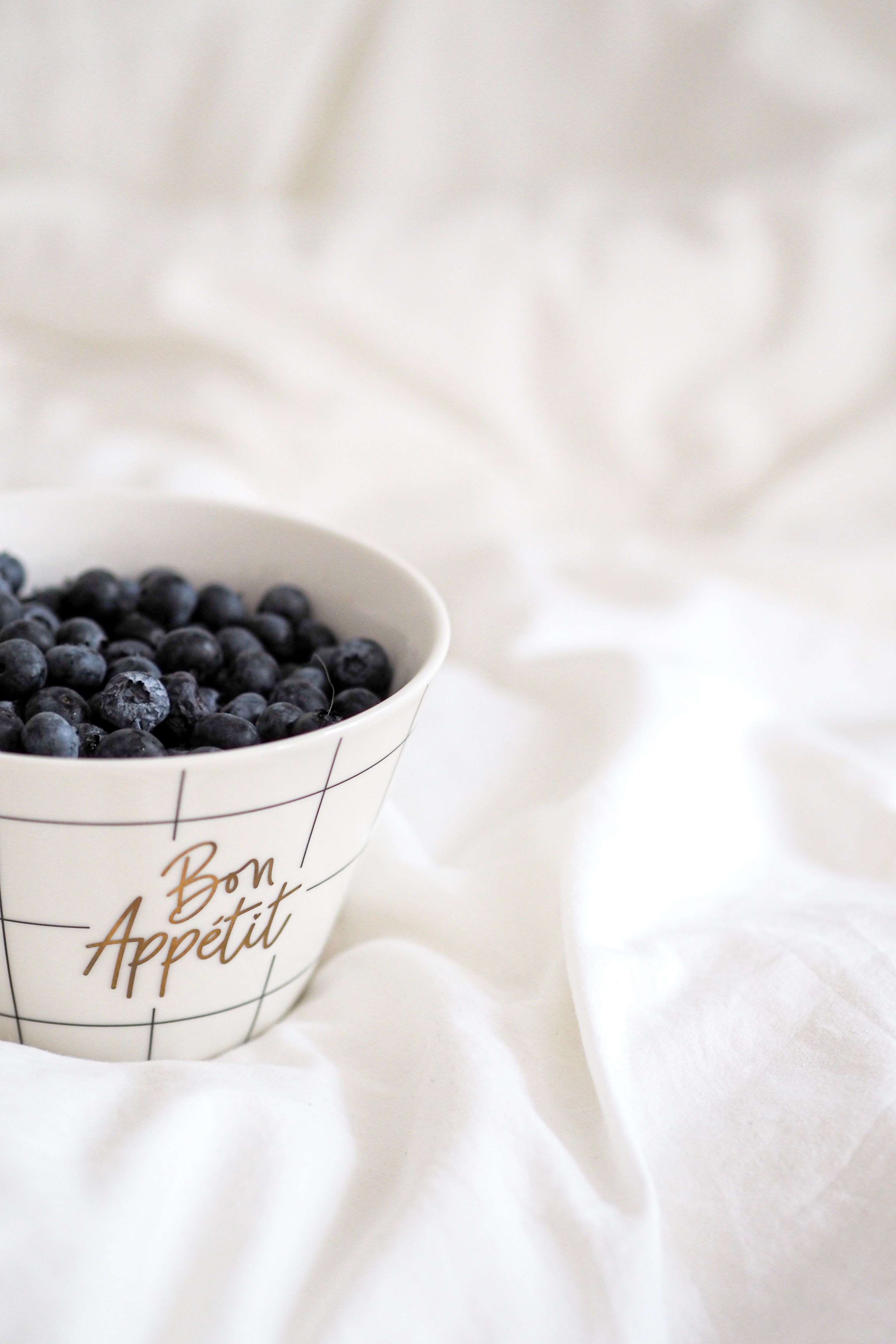 A bowl of ripe blueberries on soft white bedding, highlighting freshness and elegance.