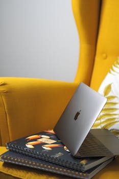 Modern workspace featuring a sleek laptop on colorful notebooks in a cozy yellow armchair.