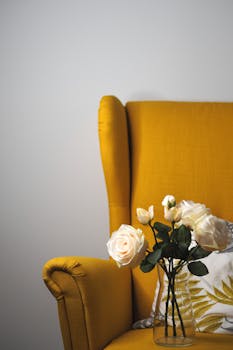 A stylish yellow armchair with a vase of white roses and a decorative pillow on a white background.