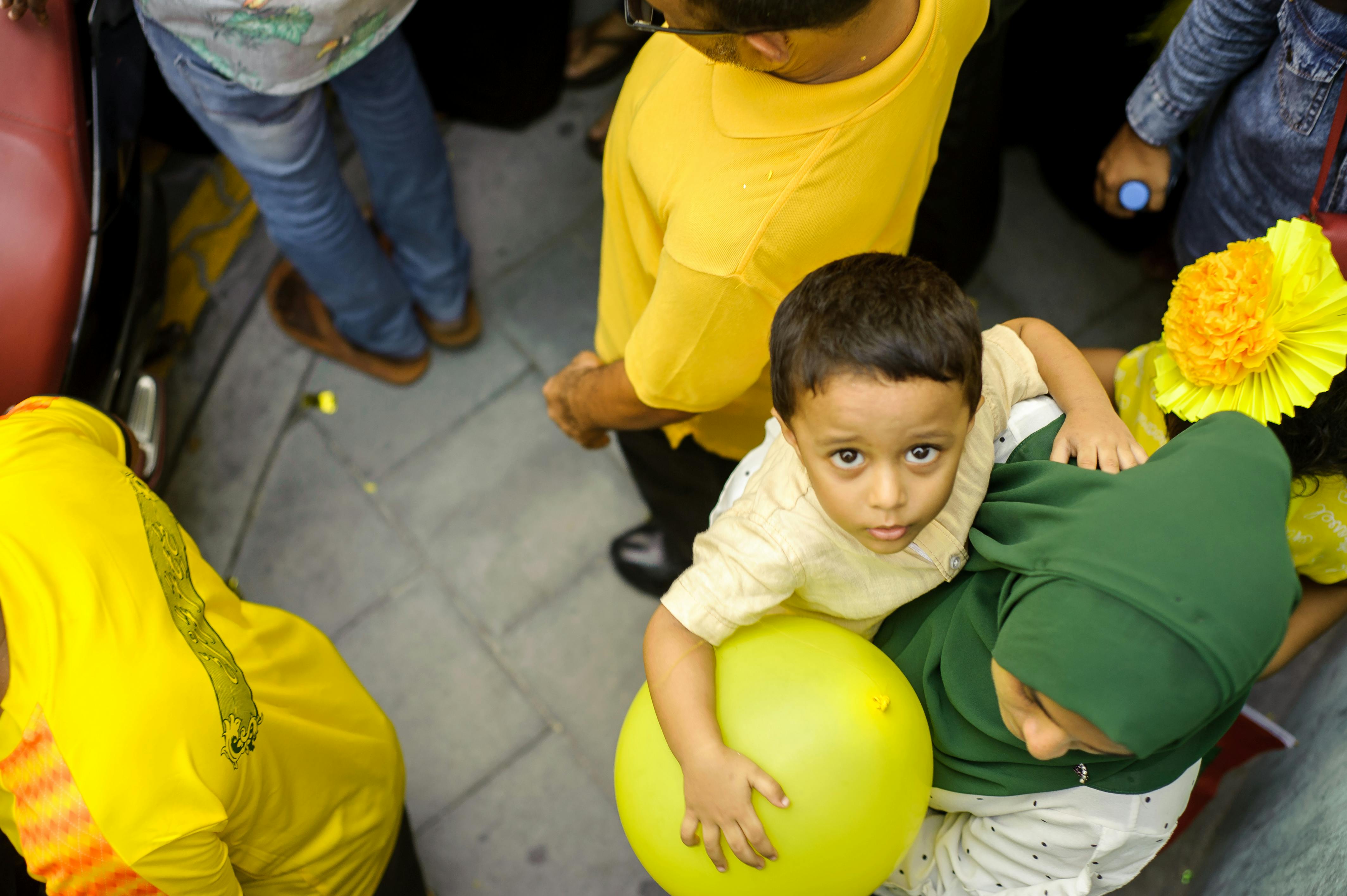 一个孩子拿着黄色气球，在繁忙的街道上抬头仰望，周围都是人