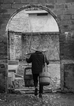 一张黑白照片中，一个人提着水桶穿过古老的拱门