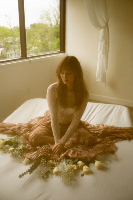 A woman in soft light sits on a bed adorned with white roses in a tranquil setting.